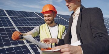 Low angle of young engineer and businessman in hardhats discussing project details against solar energy station