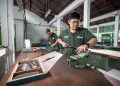 Two workers in a well-lit Indonesian workshop using clamping tools and safety gear for metalwork.