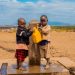 Two children filling a container at a rural water tap on a sunny day.