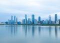 A serene view of modern skyscrapers reflecting on calm water under a cloudy sky.