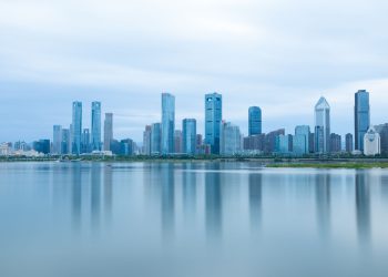 A serene view of modern skyscrapers reflecting on calm water under a cloudy sky.