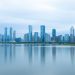 A serene view of modern skyscrapers reflecting on calm water under a cloudy sky.