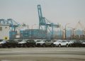 A lineup of parked cars at a bustling industrial shipping port with cranes and containers.