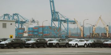 A lineup of parked cars at a bustling industrial shipping port with cranes and containers.