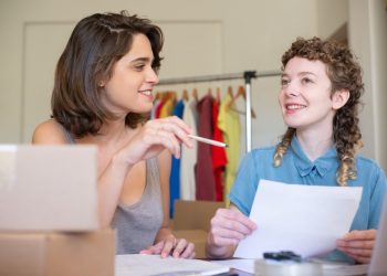 Two businesswomen discussing ideas in an office setting with clothing in the background.