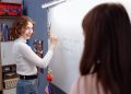 Smiling teacher writing on a whiteboard in a lively classroom setting with students.