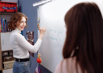 Smiling teacher writing on a whiteboard in a lively classroom setting with students.