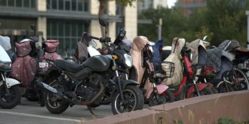 A row of parked scooters and motorcycles in an urban outdoor setting with protective covers.