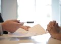 Side view of faceless formal man giving pen and paper to focused female with clenched hands at table on meeting