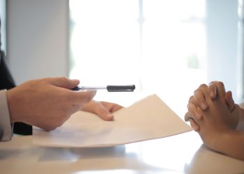 Side view of faceless formal man giving pen and paper to focused female with clenched hands at table on meeting