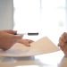 Side view of faceless formal man giving pen and paper to focused female with clenched hands at table on meeting
