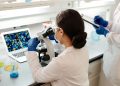 A female scientist in a lab coat examines samples under a microscope for research.