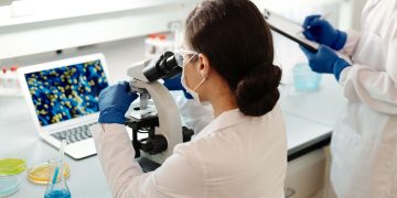 A female scientist in a lab coat examines samples under a microscope for research.