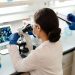 A female scientist in a lab coat examines samples under a microscope for research.