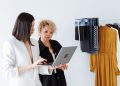 Two women fashion designers collaborating with a laptop in a modern workspace with clothing samples.