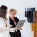 Two women fashion designers collaborating with a laptop in a modern workspace with clothing samples.