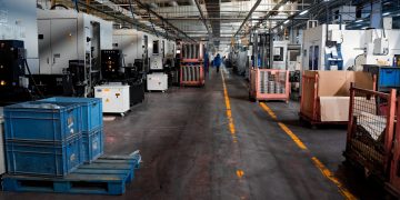 Vast industrial factory floor with machinery, crates, and structured workstations.