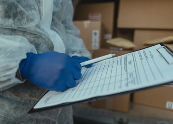 A courier wearing protective equipment writes on a clipboard among packages.