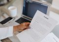 Close-up of a person holding a contract document next to a laptop during a work session.