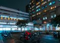 Night view of a hospital with scooters parked outside, illuminated by bright city lights.