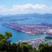 Aerial image of a bustling industrial port with containers in a scenic coastal setting.