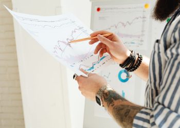 A man analyzing stock market charts with a pen, holding a paper report indoors.