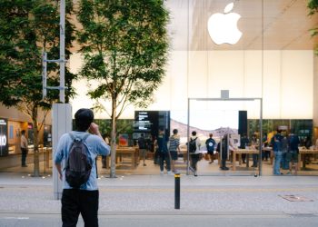 A busy Apple Store entrance with glass facade, trees, and people milling around.