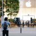 A busy Apple Store entrance with glass facade, trees, and people milling around.