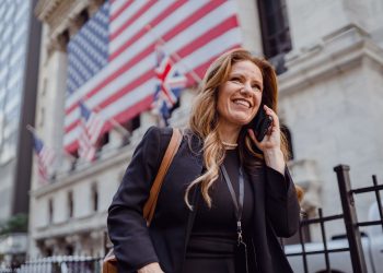 Smiling businesswoman using phone outside the stock exchange with flags in background.