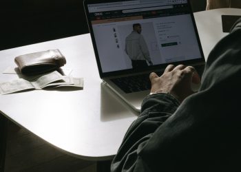 Adult man shopping for clothes online at home using a laptop, with a wallet on the table.