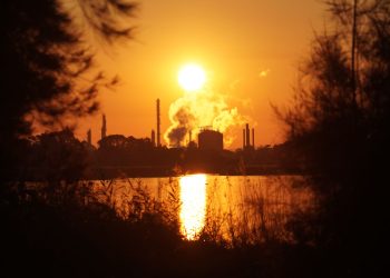 Sunrise over an industrial area reflected on water with silhouette of trees.