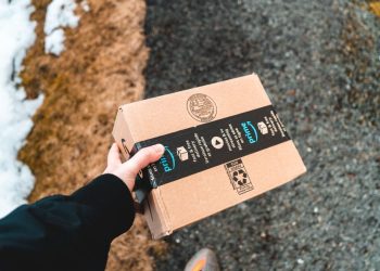 A person holding a cardboard Amazon Prime package on a snowy urban sidewalk.