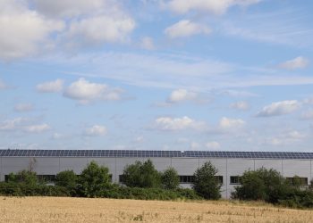 A countryside warehouse with solar panels and fields under a blue sky with clouds.