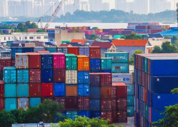 Vibrant stacked cargo containers at a bustling port with urban skyline in background.