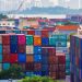 Vibrant stacked cargo containers at a bustling port with urban skyline in background.