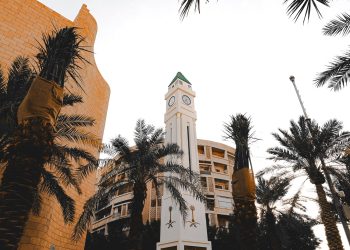 A striking clock tower surrounded by palm trees against a bright sky in an urban environment.