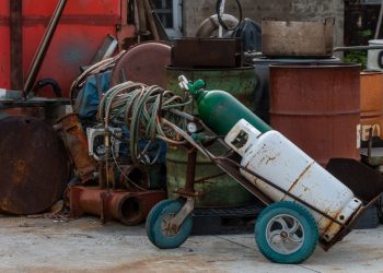 A cluttered industrial junkyard featuring gas cylinders, rusty drums, and tangled hoses.