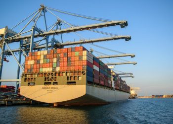 A large container ship at an industrial harbor with cranes under a clear blue sky.