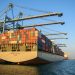 A large container ship at an industrial harbor with cranes under a clear blue sky.