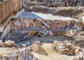 An excavator operates in a large stone quarry with rugged rocky terrain under daylight.