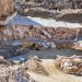 An excavator operates in a large stone quarry with rugged rocky terrain under daylight.