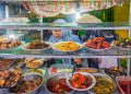 Colorful Indonesian street food stall with a variety of traditional dishes and a vendor serving customers.