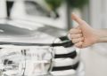 Close-up of a thumbs up gesture next to a car headlight, symbolizing approval or satisfaction.