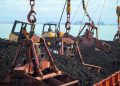 Heavy machinery loading coal at a port with a clear sky reflecting on the water.