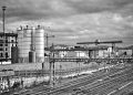 Black and white urban scene depicting a railway yard and industrial buildings under an overcast sky.