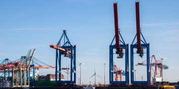 Cranes at the busy Hamburg harbor with blue skies and wind turbines.