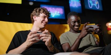Two happy young men sitting together enjoying mobile gaming indoors.